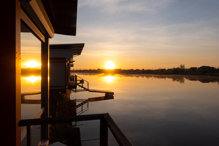 Beautiful orange sunrise over the river. Golden twilight, sunlight reflected in water ripples. Dark silhouette of the trees, rays of sunset reflected in the windows. Vacation, tranquility.の写真素材