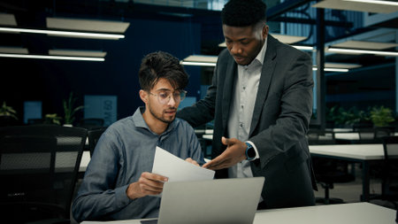 Male African boss businessman mentor teaching indian man trainee new employee discuss online business project on laptop explain details documents to colleague men working together teamwork in officeの写真素材