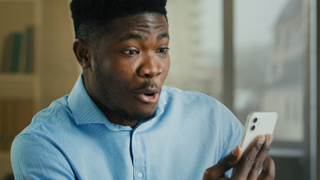 Surprised happy adult ethnic man African American businessman office worker employer business salesman use mobile phone cellphone talking virtual webcam chat say wow shock win online prize good offerの写真素材