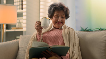 Happy smiling inspired Caucasian old woman covered with blanket on sofa drinking tea coffee with cup reading book smile atmospheric peace senior lady elderly female retired read drink at home couchの写真素材