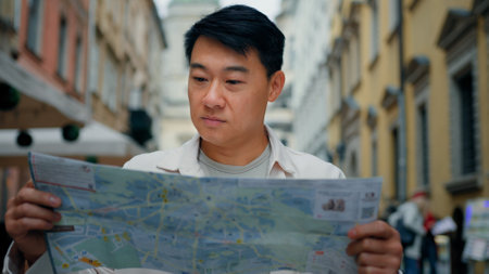 Asian male tourist traveling in new city standing outdoors looking at paper map in search of location using travel guide to get directions young man traveler international student searching right wayの写真素材