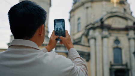 Back view unrecognizable man tourist travels in Europe photographing landmark using camera of mobile phone male blogger traveler holding smartphone taking pictures historical building in city centerの写真素材