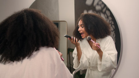 Beautiful African American woman ethnic girl in bath home looking at mirror reflection apply powder for face make up preparing to dating with cosmetics answer call angry mad screaming in mobile phoneの写真素材