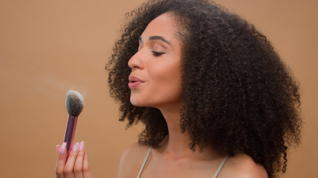 Close up portrait African American ethnic woman model girl holding brush blow away dust apply skin tone powder to nose smiling at camera at beige studio background beautiful lady cosmetics make-upの写真素材