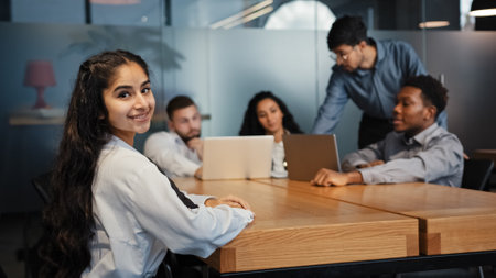 Indian ethnic businesswoman looking at camera posing in office on blurred background of brainstorming diverse office workers multiethnic multiracial coworkers discussing work with laptops at tableの写真素材