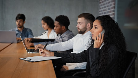 Multiracial employees sitting in conference room coworking work at table. Hispanic businesswoman CEO manager talking on mobile phone writing notes diverse multiethnic colleagues working with laptopsの写真素材