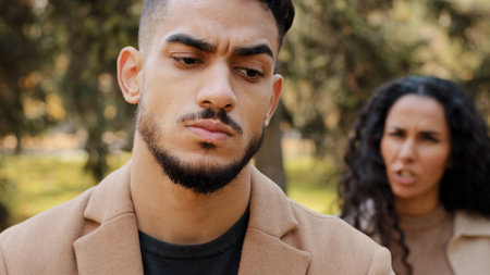 Emotional quarrel of hispanic couple outdoors angry girlfriend in background yelling and scolding boyfriend upset bearded man in foreground not listening reproaches unhappy marriage of young peopleの写真素材