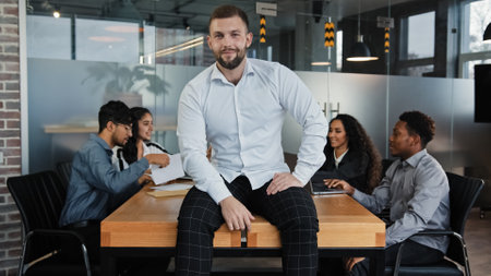 Satisfied man male leader boss company CEO businessman sitting on table desk in office smile to camera on background of multiracial coworkers colleagues teamwork project startup analyze work meetingの写真素材