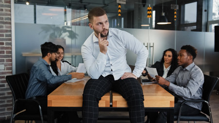 Thoughtful pensive man leader boss businessman company CEO sitting on office table thinks about solving problem difficult decision considering project idea multiracial coworkers meeting on backgroundの写真素材