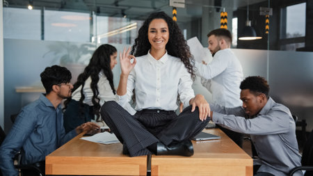 Happy calm smiling businesswoman female leader woman sitting on table in lotus position meditating in office showing ok hand gesture okay sign on background of angry colleagues team throw documentsの写真素材
