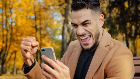 Excited hispanic guy reading good news on smartphone sitting on bench in autumn park young man happily surprised to receive message overjoyed male winner celebrating victory holding telephone outdoorsの写真素材