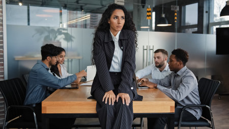 Worried sad upset leader woman worker businesswoman sitting on table in conference room thinking about business problem multicultural team colleagues in background discussing work project teamworkの写真素材