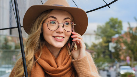 Attractive young woman with umbrella answers call on the street during rain, smart girl communicates enthusiastically, emotional caucasian lady talking on cell phone telling good news about weatherの写真素材