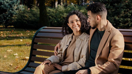 Cheerful hispanic couple cutely communicate sitting on bench in autumn city park young bearded guy hugs attractive curly girl carefree communication outdoors woman point finger spouses happily smilingの写真素材