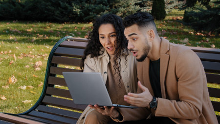 Excited hispanic couple reading good news on laptop sitting on bench in autumn park successful partners happy hugging surprised to receive email man winner celebrating victory lottery embracing womanの写真素材