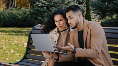 Worried hispanic young couple with laptop sit on bench in autumn park unsuccessful online shopping...