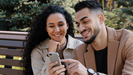 Bearded guy with attractive girl in autumn park sitting on bench cheerful couple interestedly looks at smartphone screen young man actively communicate with girlfriend partners discuss online shoppingの写真素材