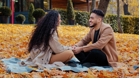 Happy cheerful hispanic couple on date in autumn park young holding hands laughing and smiling sit cross-legged on plaid in colorful leaves outdoors bearded guy emotionally talk with curly girlfriendの写真素材