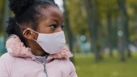 Close up African American small girl ethnic baby pupil schoolgirl ill sick daughter child kid in medical face mask looking away to side outdoors in city back to school in pandemicの写真素材