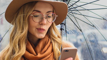 Portrait of pensive thinking puzzled confused young girl student with phone stand in street with umbrella Caucasian woman in hat and glasses in city uses mobile app female looking at smartphone chatの写真素材