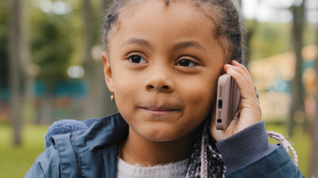 Little ethnic girl talking on mobile phone outdoors portrait funny multiracial child answer telephone call small African American baby kid daughter schoolgirl standing in street talk with smartphoneの写真素材