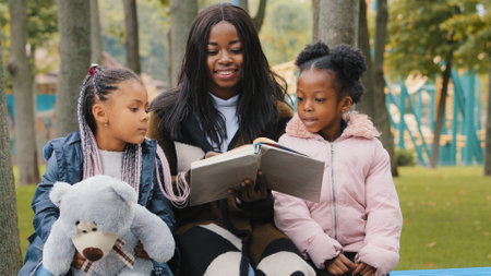 Happy african american family resting on bench in park young mother reads book to daughters child holding teddy bear little girl points finger textbook mom smiling happily together spend time outdoorsの写真素材