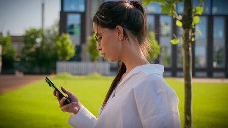 Caucasian woman businesswoman student business lady girl walk on street with green lawn using mobile phone check email browsing social network make order online store on smartphone outdoors side viewの写真素材