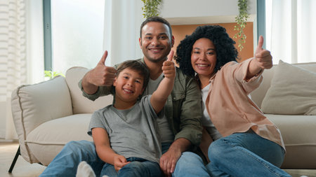 Happy multiracial family African American parents embrace with Caucasian little boy son adopted kid on home floor talk laugh fun weekend smiling father mother and cute child showing thumbs up gestureの写真素材