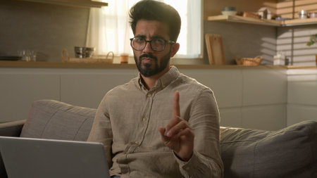 Arabian Indian man businessman freelancer working with laptop from home kitchen dissatisfied guy on sofa waving index finger negative warn sign no never warning gesture caution forbid disagree rejectの写真素材