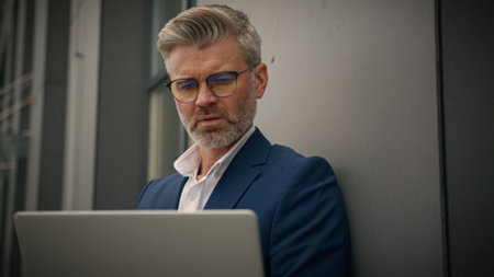 Caucasian elderly senior man mature business manager busy middle-aged employer businessman leaning wall office building typing laptop working computer exhale tired overworked take off glasses outdoorsの写真素材