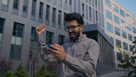 Excited Indian Arabian man male businessman watching video on mobile phone outdoors winning online match with smartphone amazed happy guy office employer in city celebrate achievement business successの写真素材