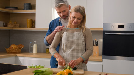 Happy family Caucasian middle-aged adult at kitchen woman cook vegetable salad cut cucumber husband man hugging cuddling embracing wife couple talking discuss diet food affectionate love talk cookingの写真素材