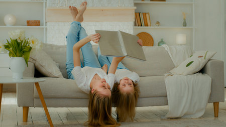 Cheerful Caucasian mother lying upside down on comfortable sofa with little child girl kid daughter reading book read together looking at pictures in textbook album family enjoy weekend at home couchの写真素材