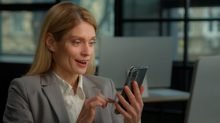 Caucasian middle-aged adult woman mature business female lady businesswoman scrolling browsing mobile phone shock wonder amazed surprise with online achievement success bet smartphone prize in officeの写真素材