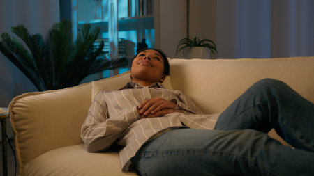 Tired African American woman in evening night living room lying on cozy sofa falling down on comfortable soft couch relaxed tranquil calm girl dream rest at home relaxing stress free weekend leisureの写真素材