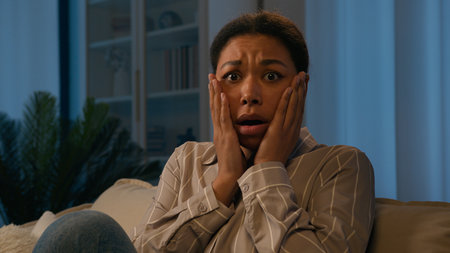 Scared African American woman frightened alone at evening home on couch shocked confused female girl feel panic suffer stress fright amazed afraid scare terrible night horror touch face shock emotionの写真素材