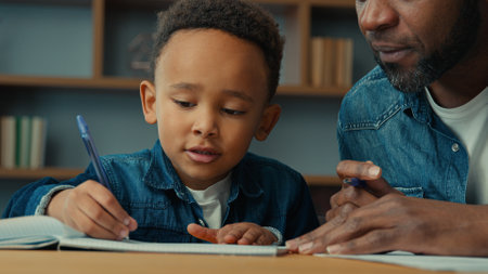 African American cute little boy child schoolboy with ethnic adult father writing homework. Dad help son with lesson class write explain exercise task kid close ears homeschooling elementary educationの写真素材