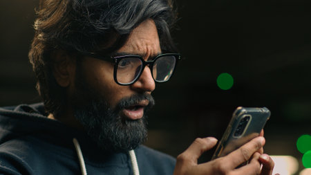 Shocked stressed annoyed disappointed upset Indian man in glasses Arabian bearded male hold mobile phone stress shock by SMS notification bad news failure business lost with smartphone at parking lotの写真素材