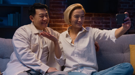 Happy excited multiracial multiethnic couple married diverse Asian man and Caucasian woman talking online video call conference with mobile phone app talk waving hello at night evening on couch sofaの写真素材