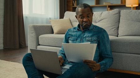 Surprised mature african american man enter data into laptop from paper documents happy with annual...