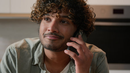 Cheerful Indian young man with curly hair talking phone in home kitchen. Happy friendly Hispanic guy talk with friends by smartphone Arabian male speaking smiling laughing pleasant mobile conversationの写真素材