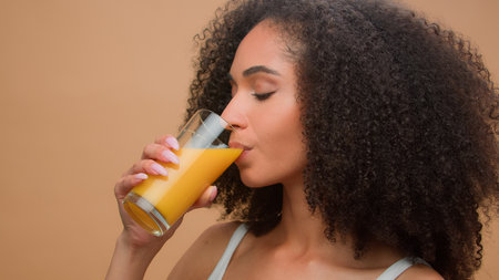 Beautiful happy healthy African American woman ethnic girl female model with curly hair drinking fresh orange juice vitamin drink diet detox organic vegetarian health care smiling in studio backgroundの写真素材