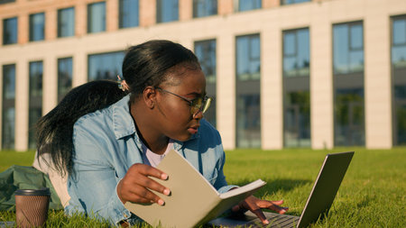African American happy woman university college academy student teen girl high school lying on green grass in park studying with book copybook laptop computer drink coffee prepare for exam test studyの写真素材