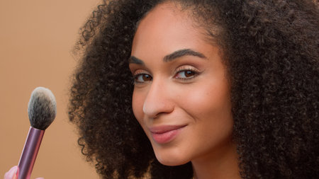 Close up portrait beige studio background beautiful African American woman with perfect skin blow to cosmetic brush apply powder at nose smiling looking at camera attractive ethnic girl make-up beautyの写真素材