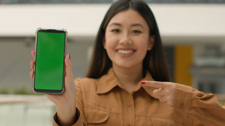 Asian chinese woman hold smartphone in front show mobile phone with green screen mockup advertisement digital app advertise cellphone web service korean girl point finger at empty chroma key displayの写真素材