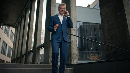 Walking outdoors going old mature Caucasian business man employer talk smartphone negotiate speaking cellphone walk in city near office company building talking mobile phone call senior businessmanの写真素材