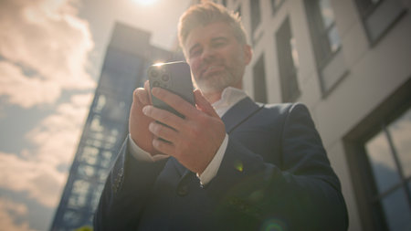 Smiling old man middle aged businessman using mobile phone outdoors in sunshine. Happy senior adult business male user employer holding cellphone texting smartphone online chat city building sunbeamの写真素材