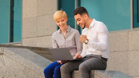 Diverse colleagues Caucasian woman and arabic Hispanic man sitting outdoors looking in laptop successful win. Businesswoman with computer makes offer to businessman winner making victory gestureの写真素材