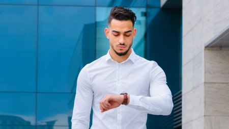 Serious upset angry hispanic indian man bearded guy disgruntled businessman boss wears formal stylish shirt stands outdoors looking at wrist watch worried about being late lateness time rush waitingの写真素材