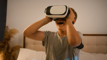 Funny enthusiastic smiling child kid boy schoolboy play video game using innovative digital VR goggles playing online 3d AR technology in metaverse world wearing virtual reality glasses at home on bedの写真素材